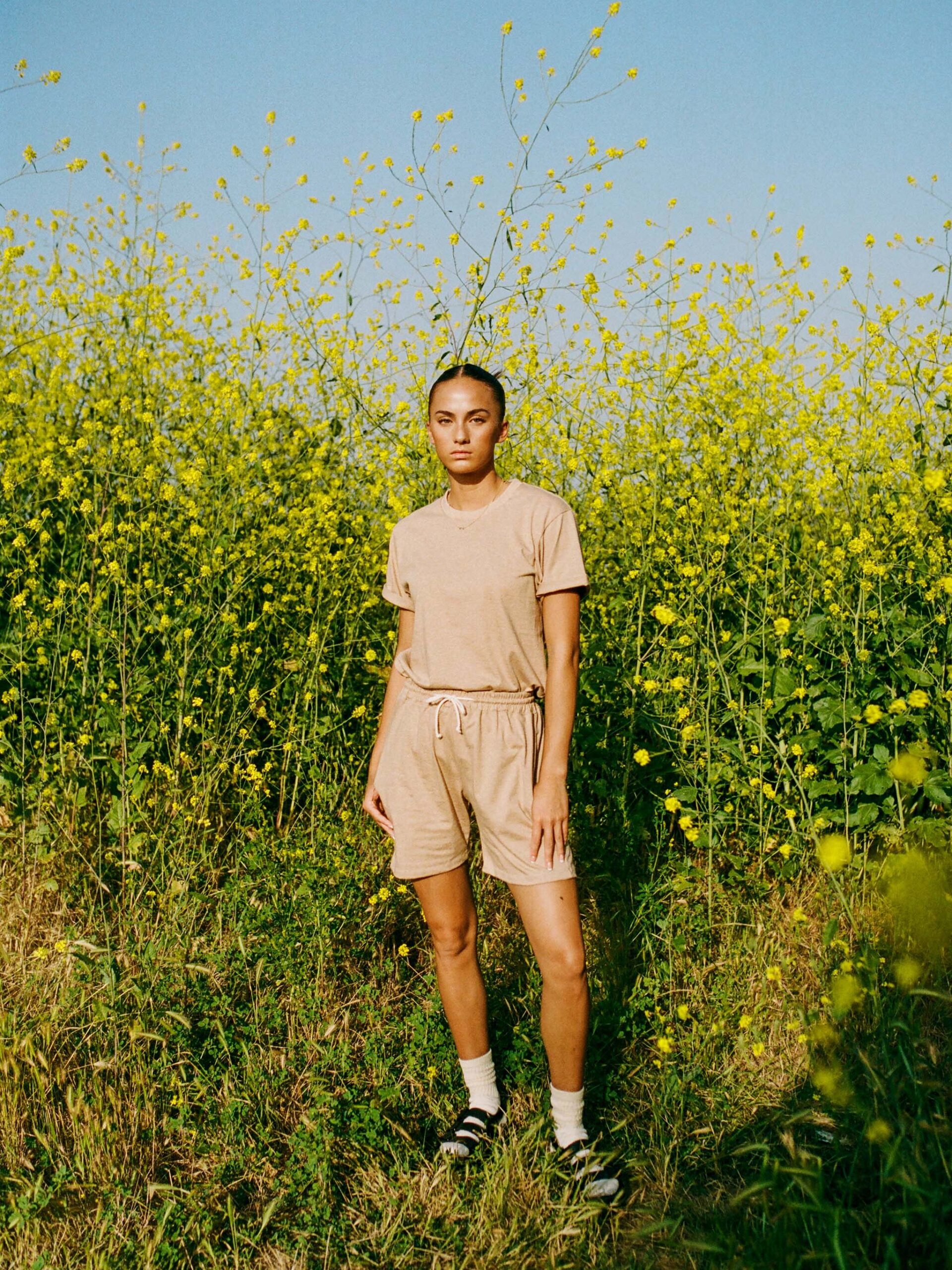 Person standing in front of tall yellow flowering plants, wearing a beige t-shirt and shorts with white socks and sandals.