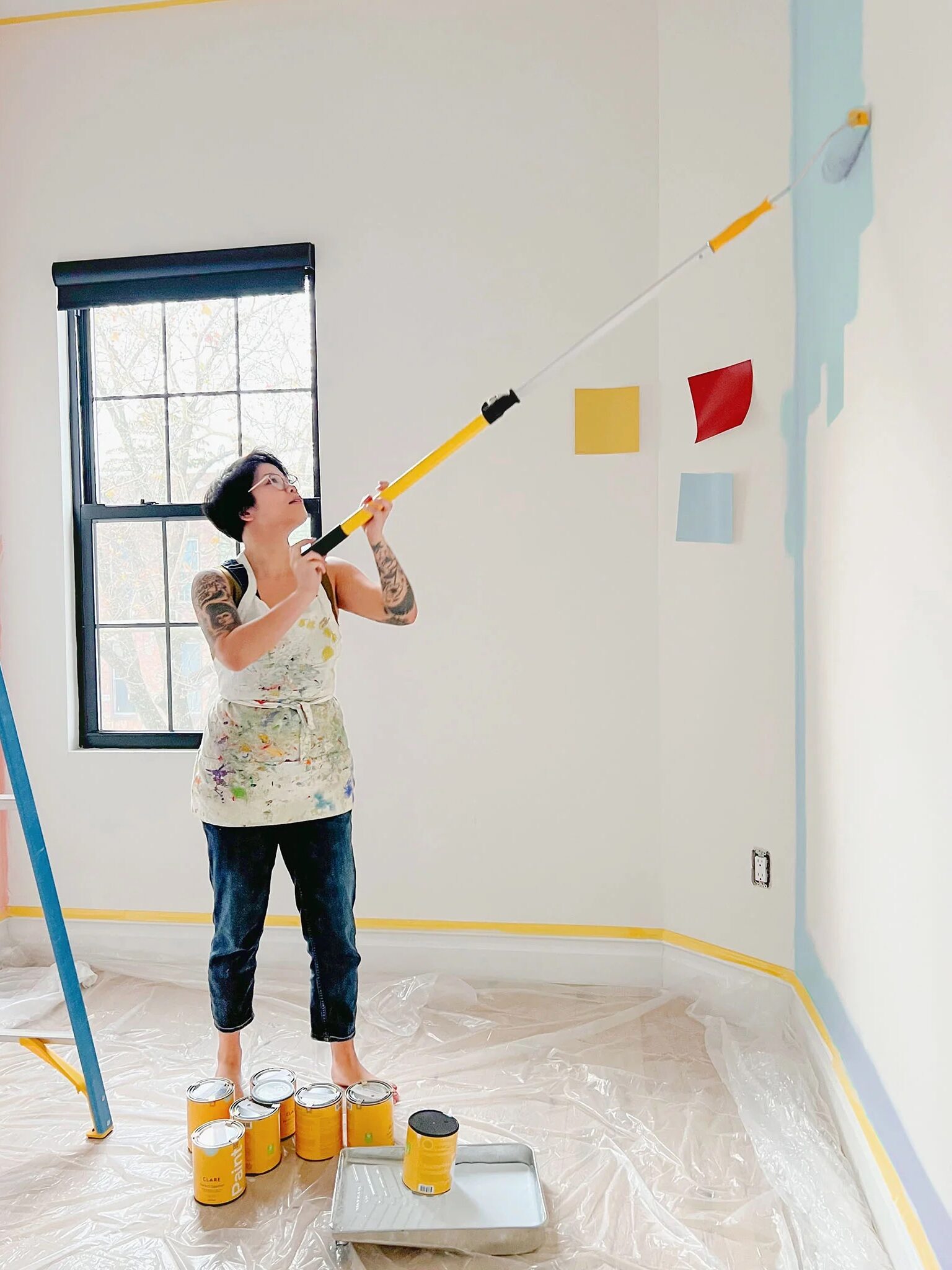 A person with tattoos stands barefoot on paint cans while using a roller brush to paint a wall light blue. A ladder and color swatches are visible nearby in a room with plastic-covered floors.