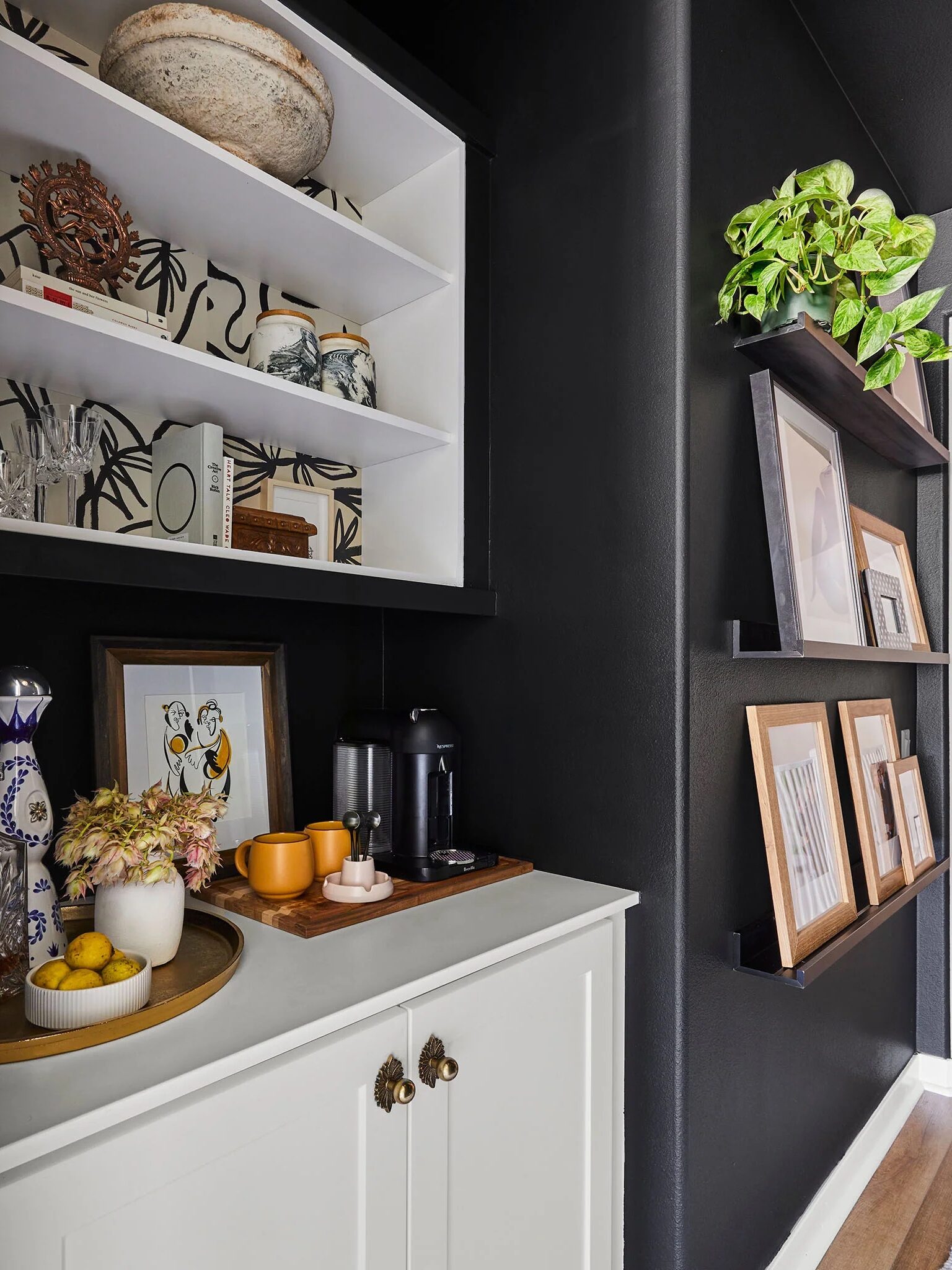 A small kitchen corner with black walls, open shelves with framed pictures and decorations, a white cabinet with a coffee maker, cups, and a tray holding lemons and a vase with flowers.