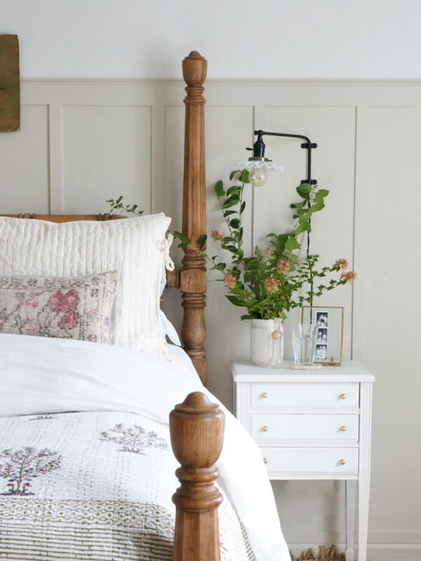 A bedroom with a wooden four-poster bed, white bedding set, and pillows. A white nightstand beside it has plants, a lamp, and framed photos. Wall art hangs above the bed.