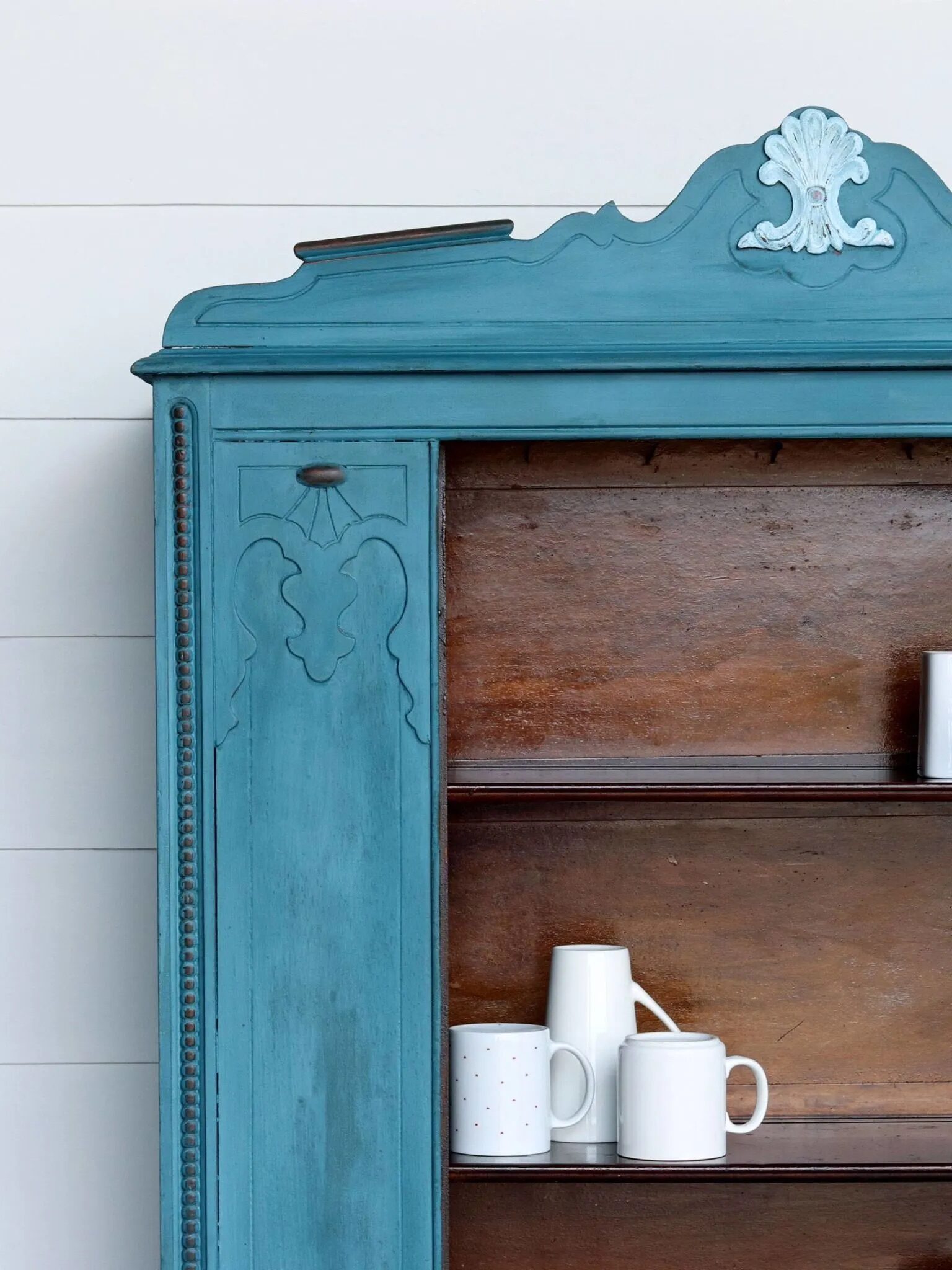 A blue wooden cabinet with detailed carvings is partially visible against a white shiplap wall. The cabinet has open shelves, with a few white cups and mugs placed on it.