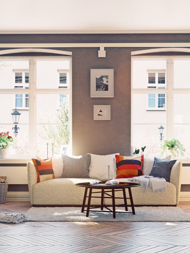 A sunlit living room features a beige sofa with colorful pillows, a round wooden coffee table, floor lamps, and large windows with light streaming in. Decor and flowers add coziness to the space.