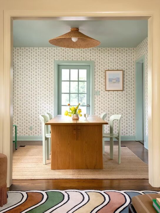 A dining room with a wooden table, light green chairs, a rattan light fixture, patterned wallpaper, an area rug, and a vase with yellow flowers.