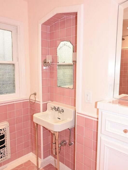 A small, vintage bathroom with pink tile walls, a white pedestal sink, and a mirror. The room also has frosted glass windows and a cabinet with a pink tile countertop.