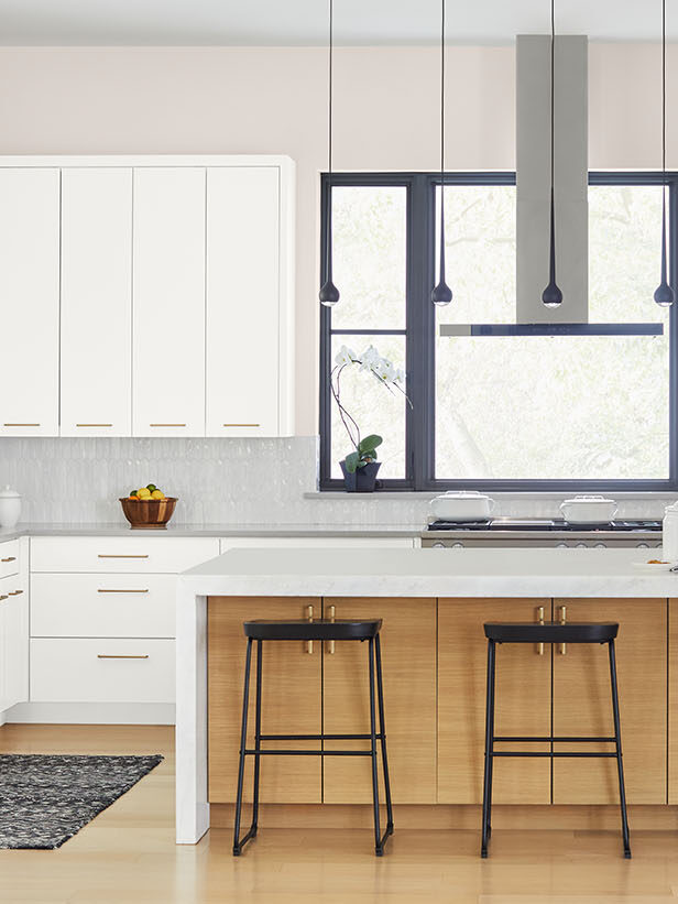 Modern kitchen with white cabinetry, black countertops, a large island with three wooden stools, stainless steel appliances, and a rug on a light wooden floor. Large windows provide natural light.