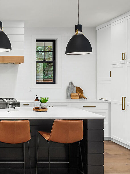 Modern kitchen with white cabinets, black island, brown leather barstools, pendant lights, and stainless steel appliances. Two windows are on the back wall above the sink and stove area.