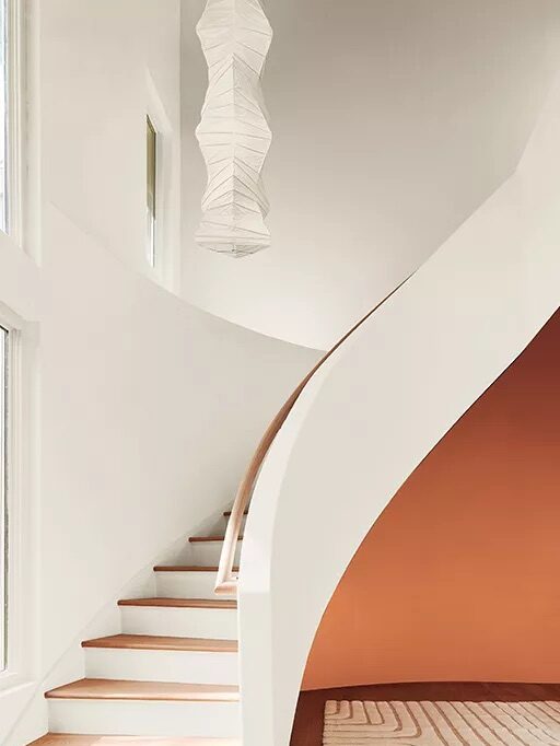 A minimalist curved staircase with wooden steps beside a white wall, leading up to a landing. A geometric light fixture hangs above, and an orange accent wall is visible along the side of the stairs.