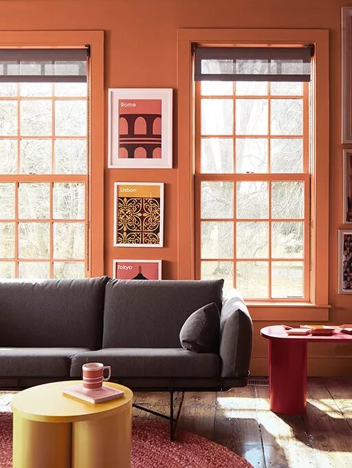 A room with orange walls, two large windows, a gray couch, and three framed pictures. There is a yellow circular table and a red side table.