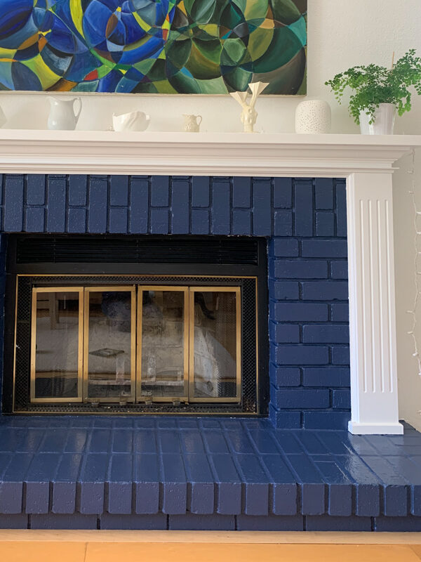 A fireplace with a blue brick surround and white mantle, featuring a modern painting, decorative items, and a small potted plant on the mantle.