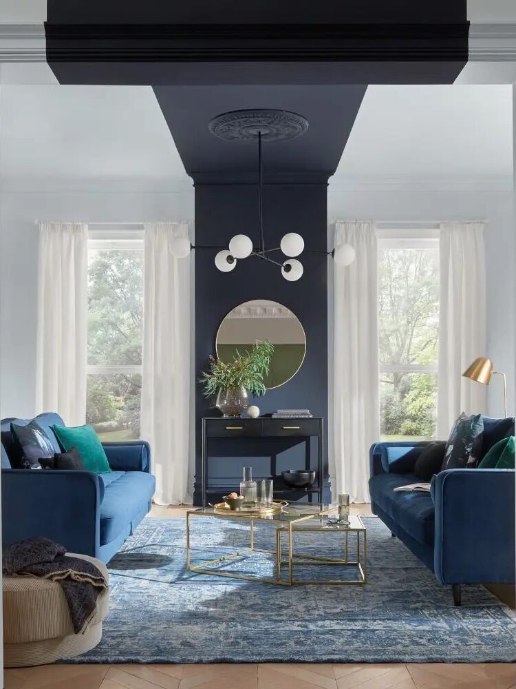 A modern living room with blue sofas, a blue area rug, gold accent tables, a central console with a round mirror, and white curtains framing large windows.