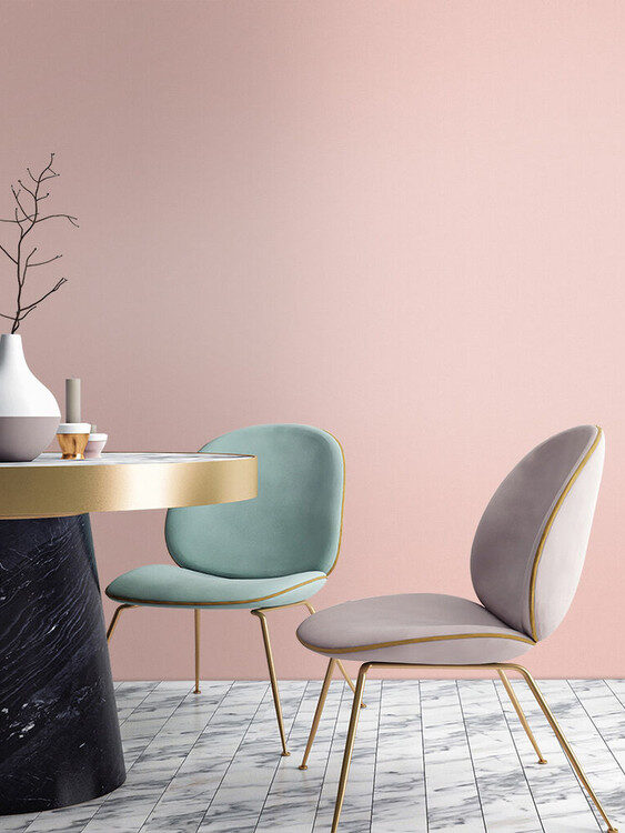 Two modern chairs with slim gold legs, one mint green and one light gray, next to a round marble table with vases, set against a pastel pink wall.