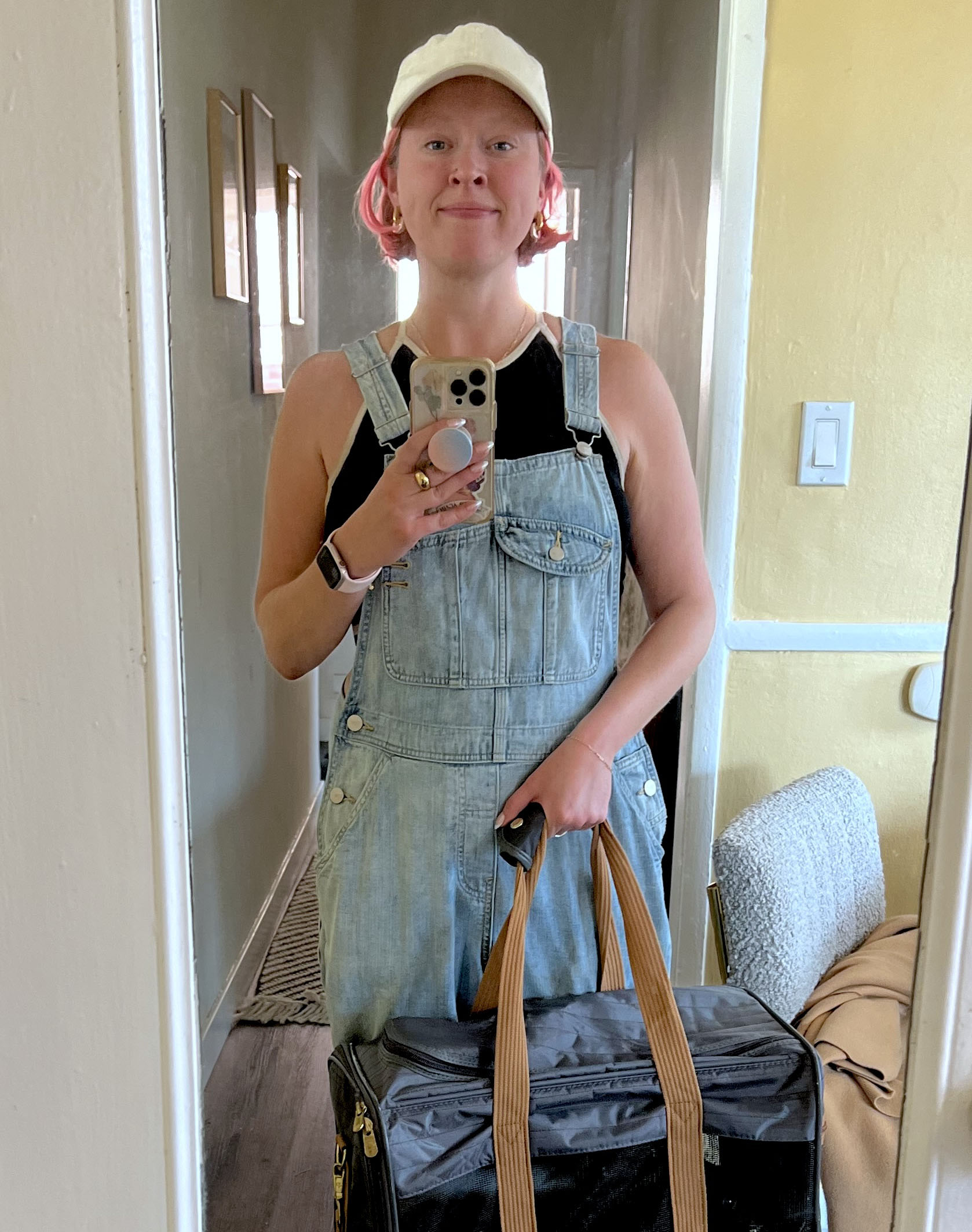 Person in overalls and a cap takes a mirror selfie, holding a black bag with tan handles.