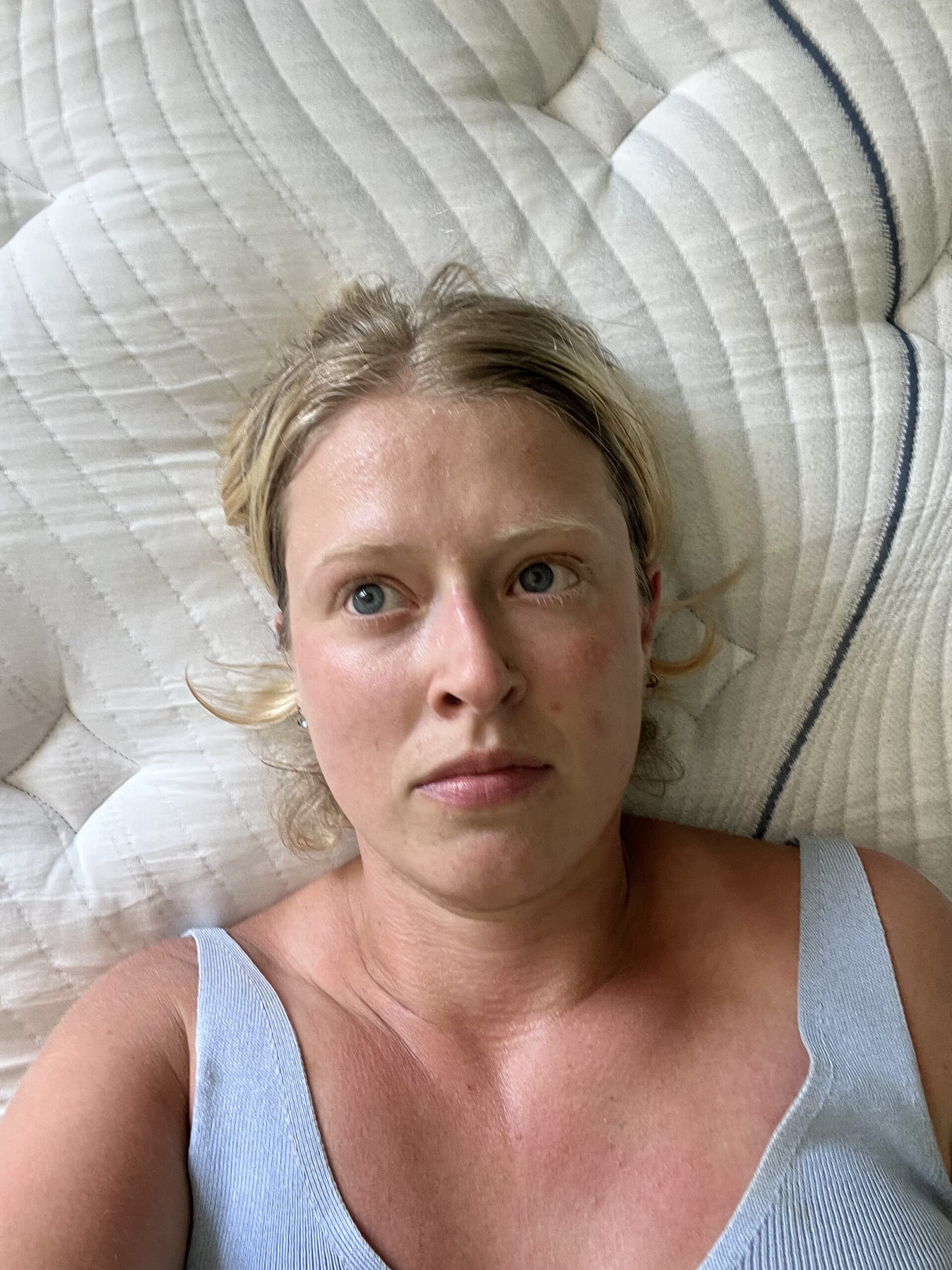 A person with light hair and a neutral expression lies on a quilted surface, wearing a light-colored sleeveless top.