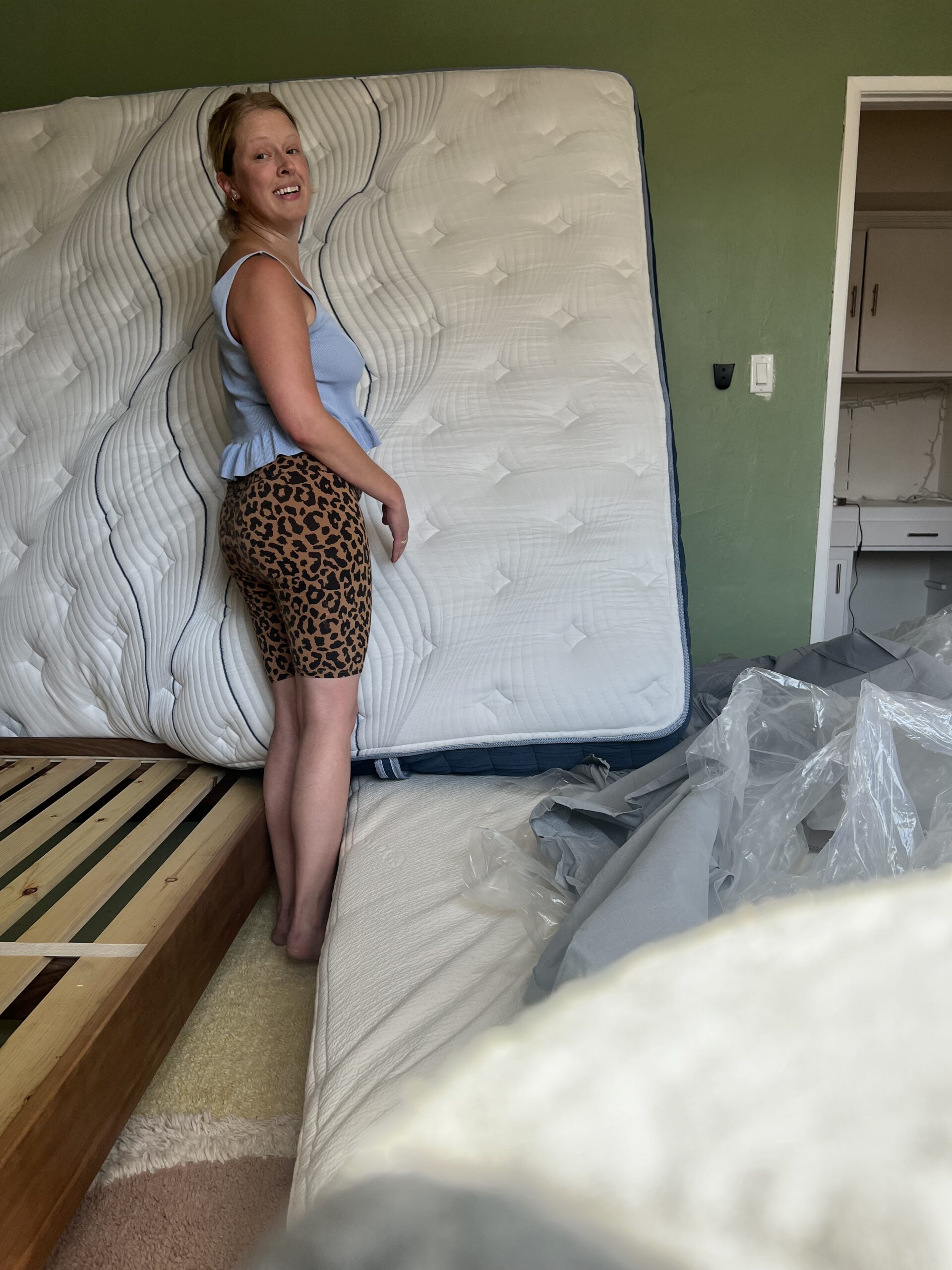 A woman stands next to an upright mattress in a bedroom. She wears a blue top and leopard print shorts. The room appears to be in the process of being furnished.