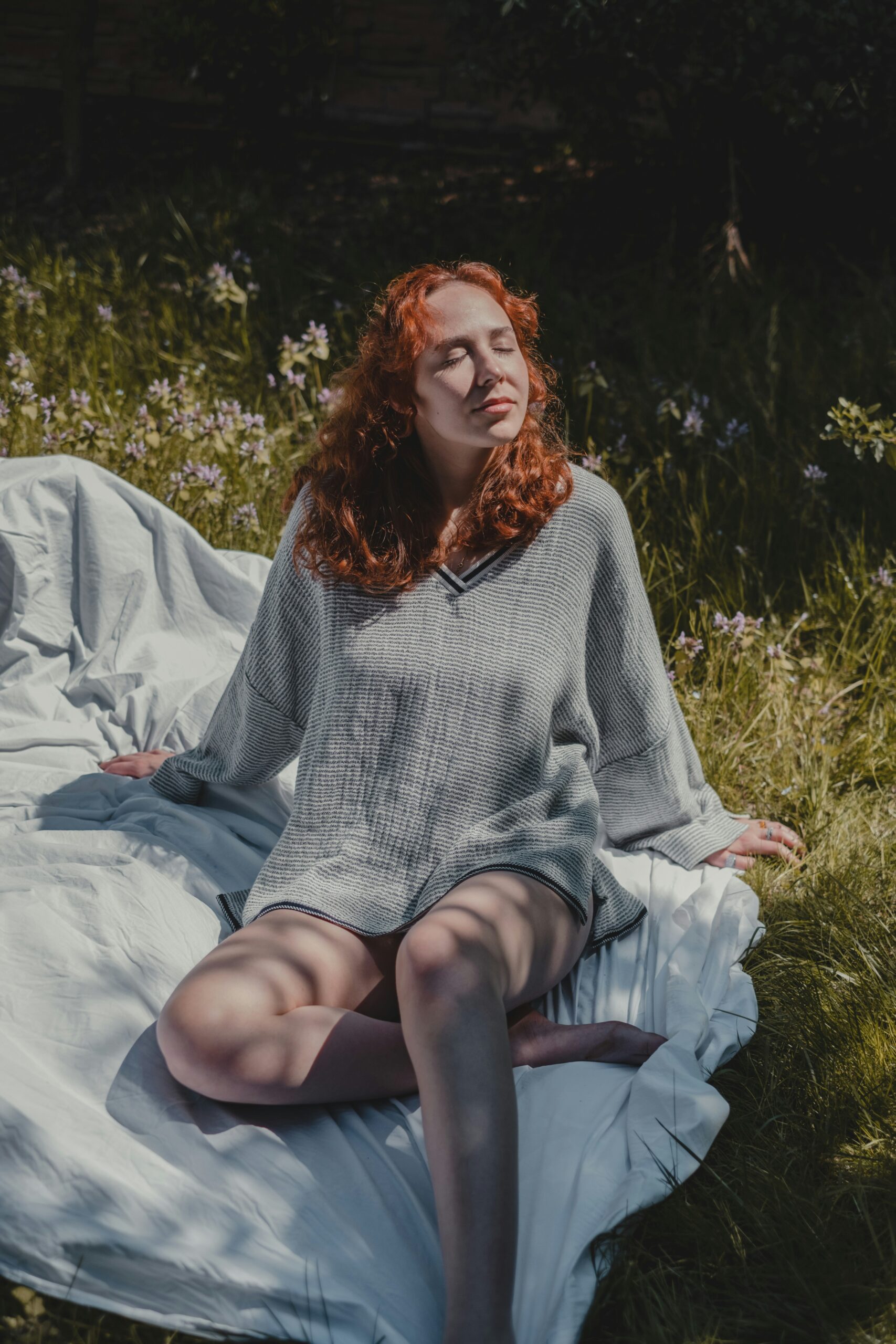 A person with red hair sits on a white sheet in a grassy area with their eyes closed, enjoying the sunlight.