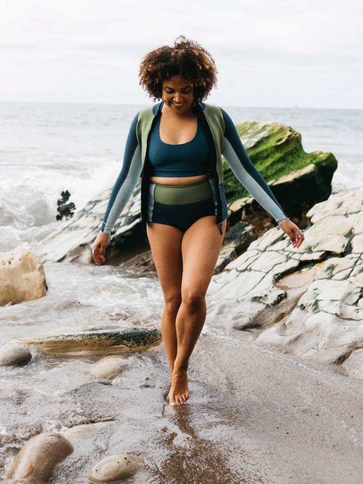 Person in a blue and green wetsuit walks barefoot on a rocky beach with the ocean in the background.