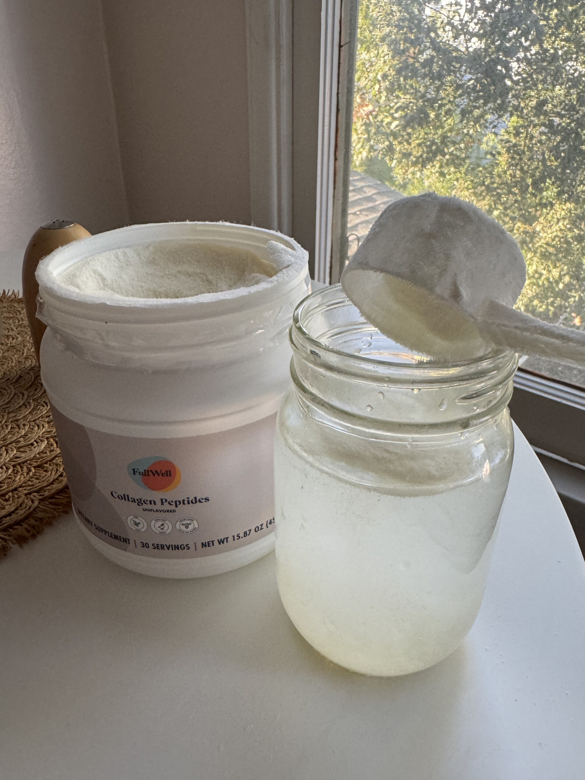 Open container of collagen peptides with a scoop being added to a glass of water, placed on a table near a window.