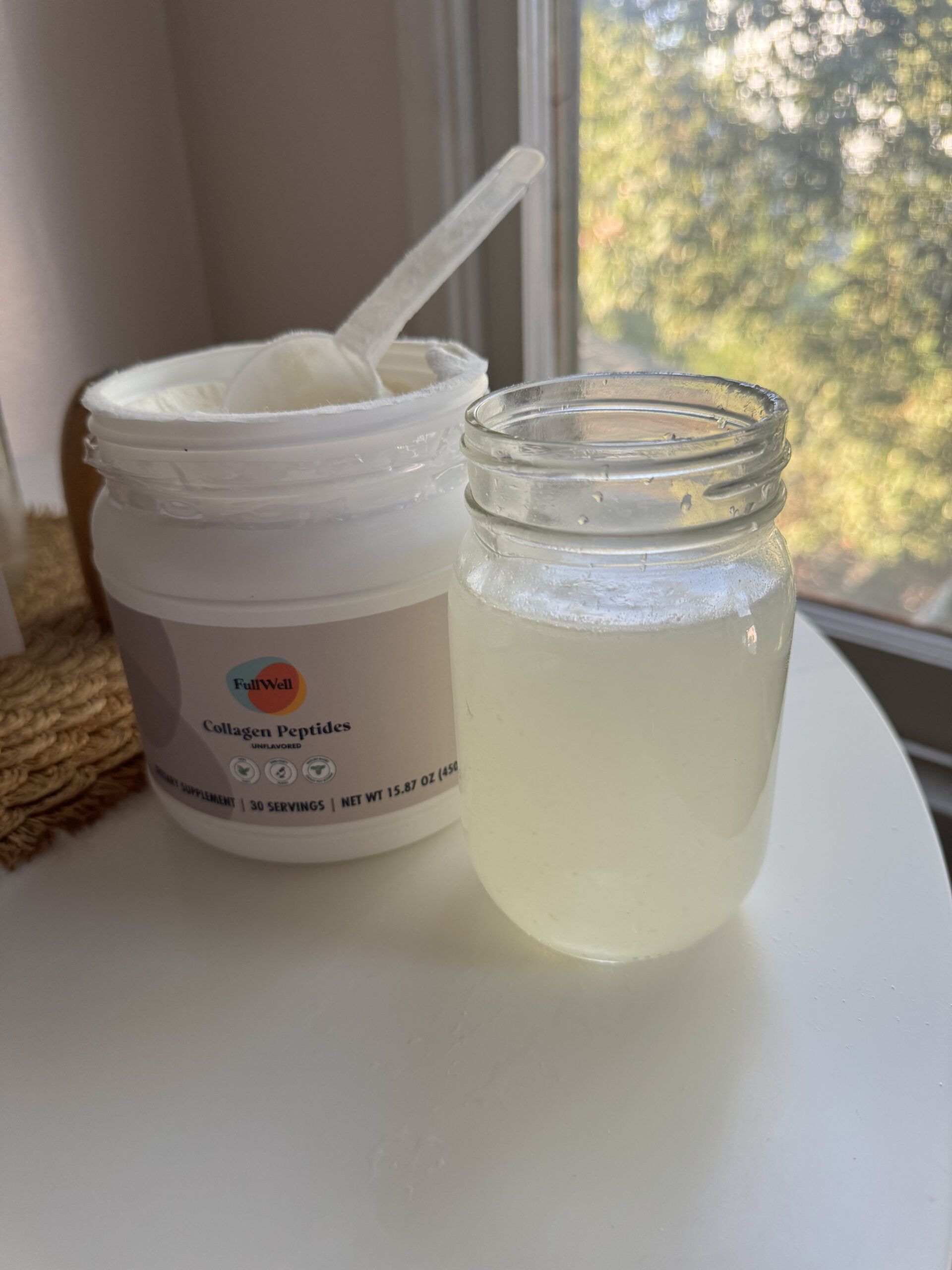 A jar of collagen peptides sits open next to a mason jar filled with a clear liquid on a white table by a window.