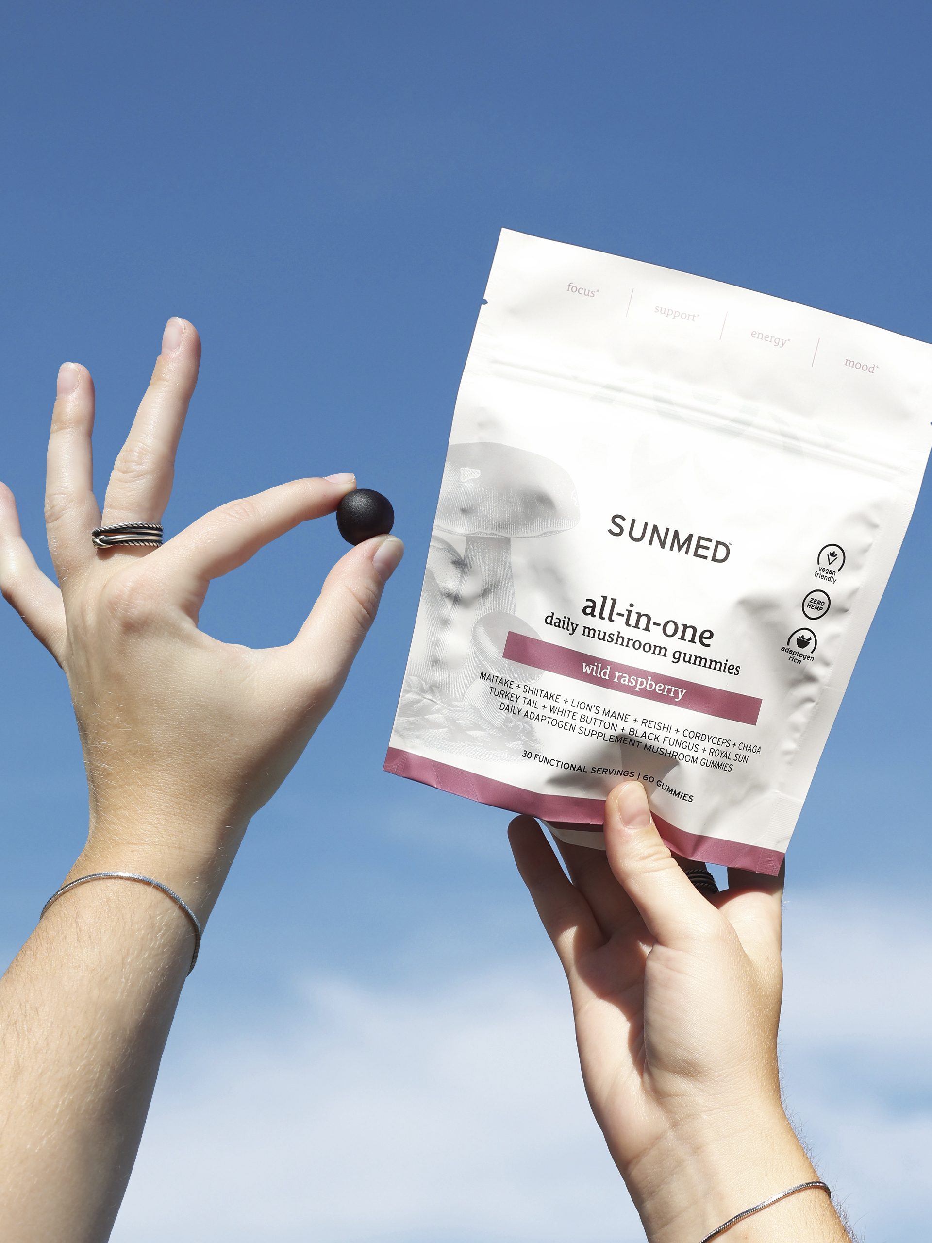 Hands holding a packet of “Sunmed all-in-one daily mushroom gummies” and a single black gummy against a clear blue sky background.