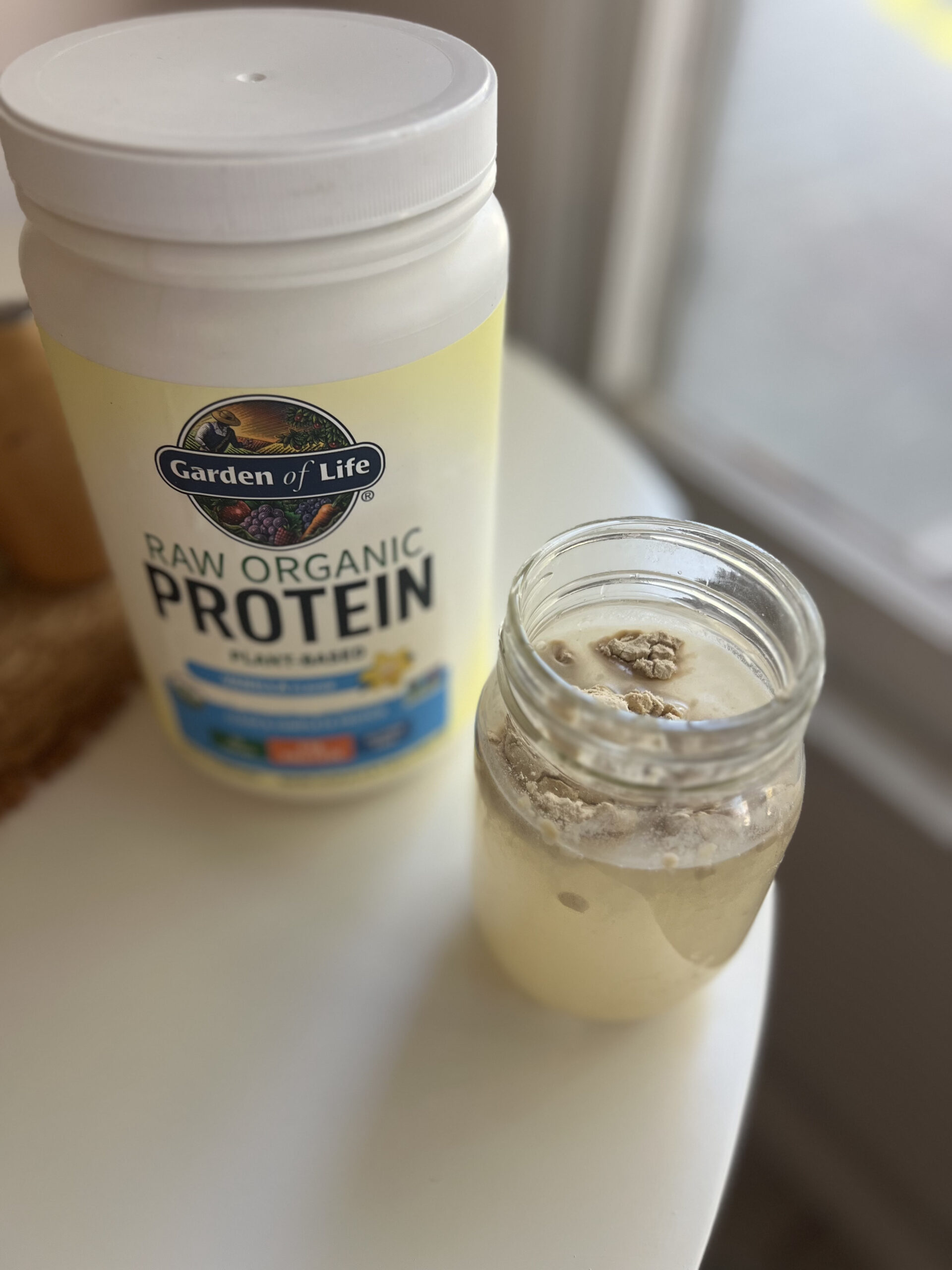 Container of Garden of Life Raw Organic Protein next to a glass jar with a protein shake mixture on a white table.