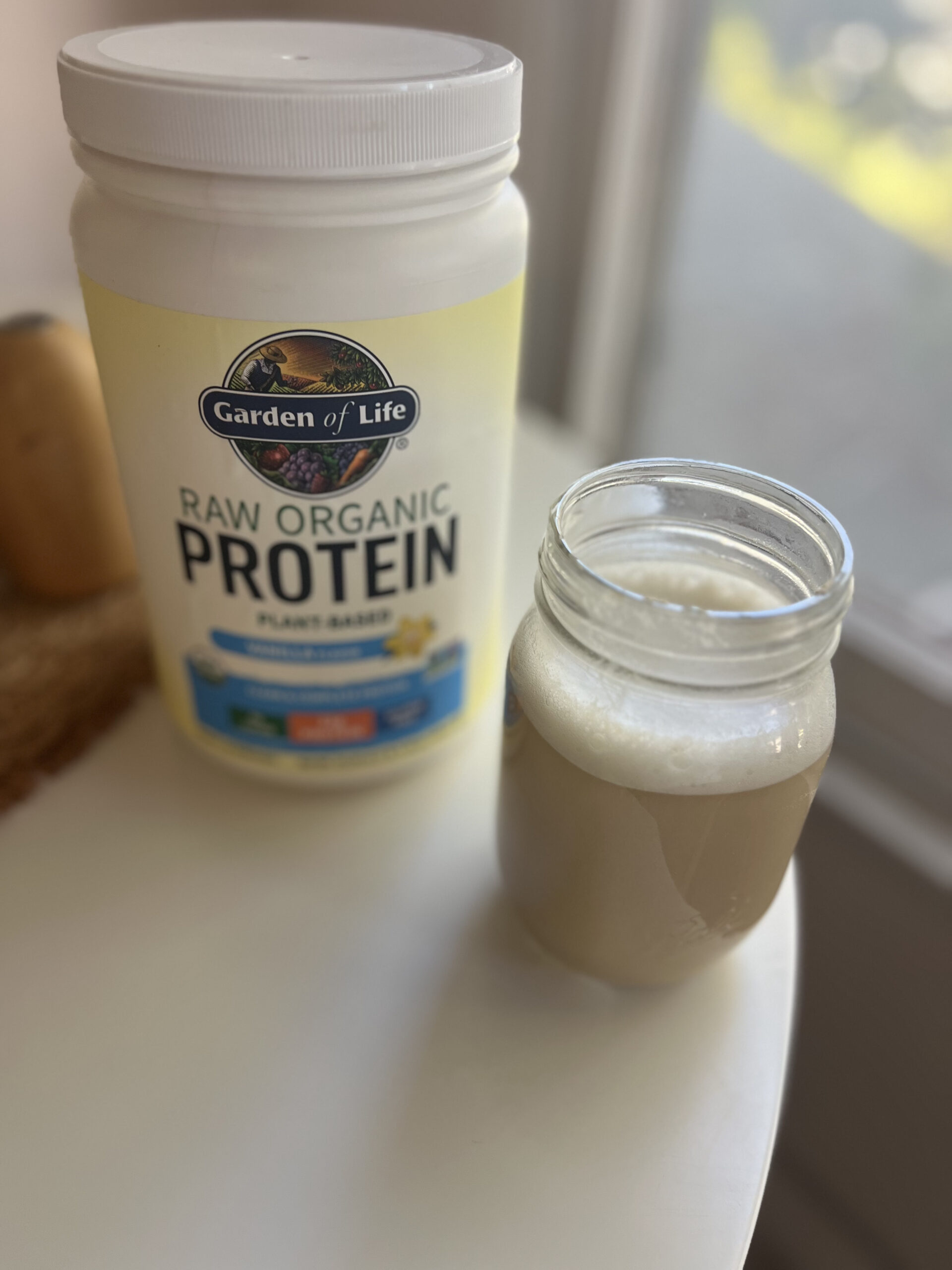 Container of Garden of Life raw organic protein powder next to a glass jar filled with a light brown protein shake on a table.