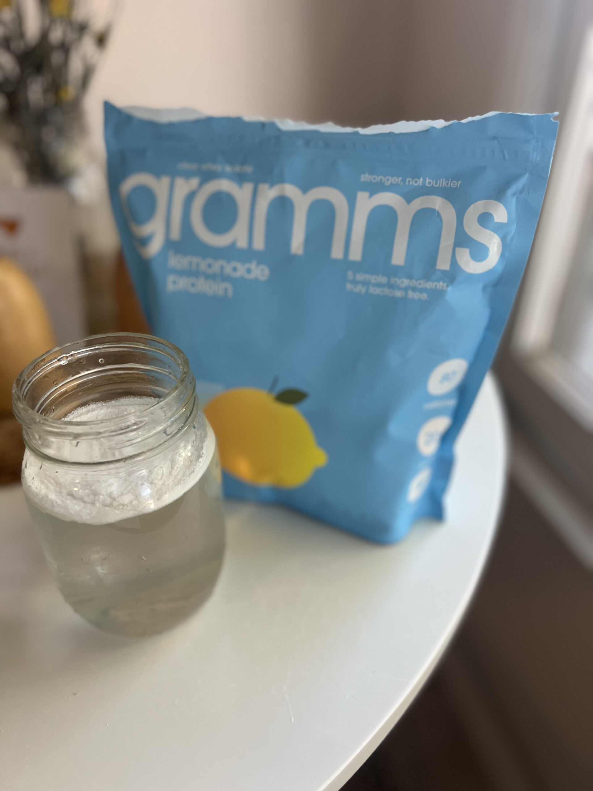 Blue bag of "gramms lemonade protein" next to a mason jar filled with a clear drink on a white table.