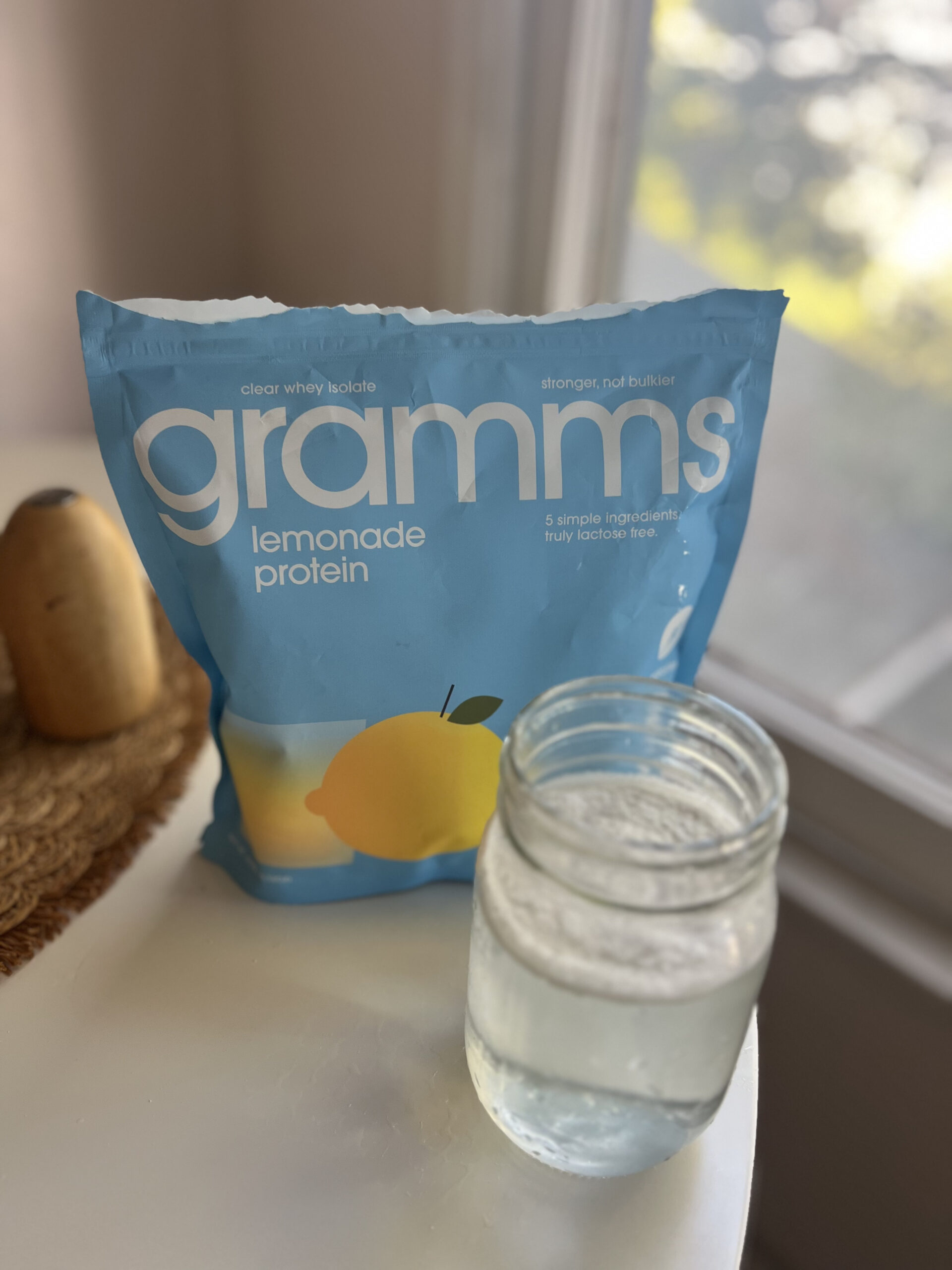 Bag of Gramms lemonade protein powder next to a glass jar filled with a clear liquid on a white table.