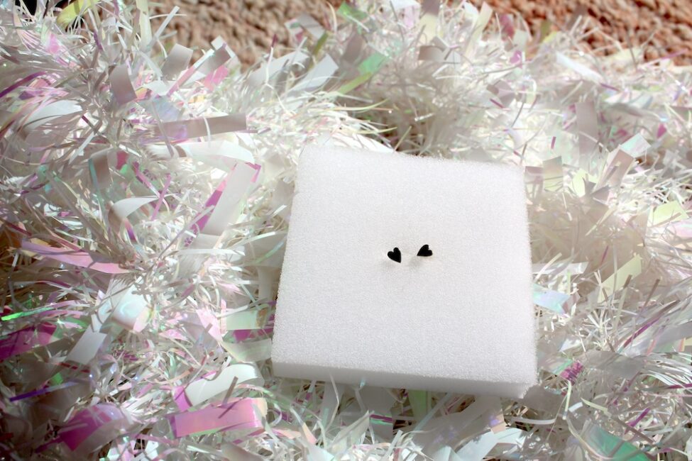 A pair of small heart-shaped earrings on a white foam block, surrounded by iridescent decorative material.