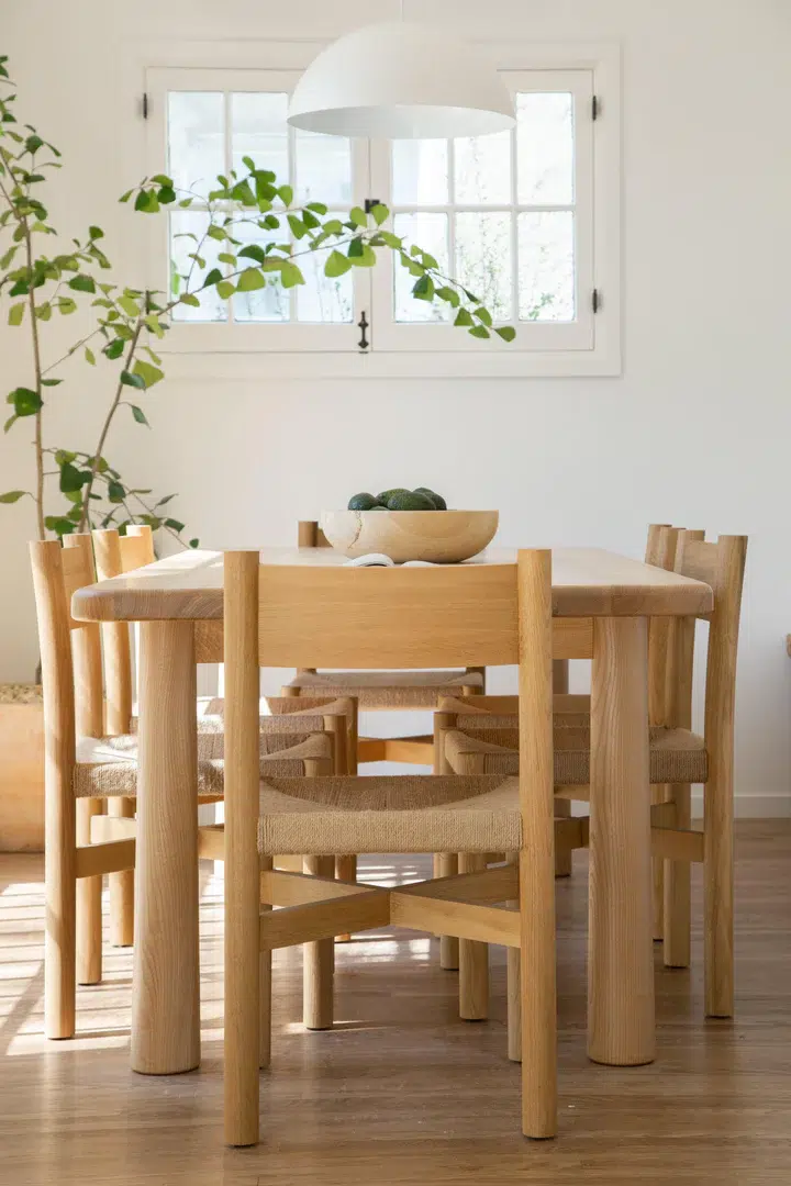 A wooden dining table with four chairs, a bowl of fruit, a hanging light fixture, and a tall plant in a bright room with wooden flooring.