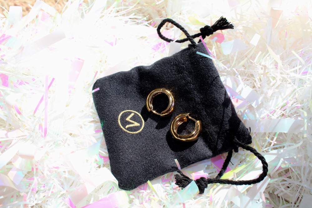 Two gold hoop earrings on a black drawstring pouch with a gold logo, placed on a bed of iridescent shredded paper.