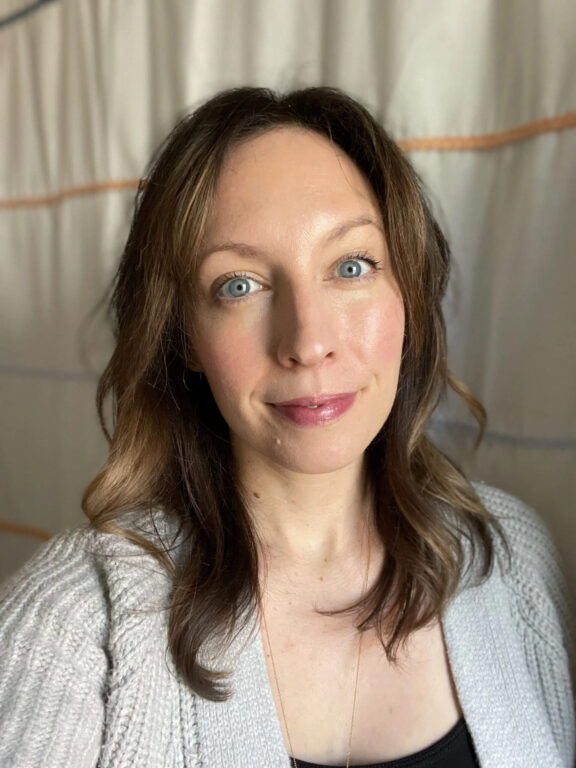 A woman with shoulder-length brown hair and blue eyes is wearing a light-colored sweater, standing in front of a light, draped backdrop.