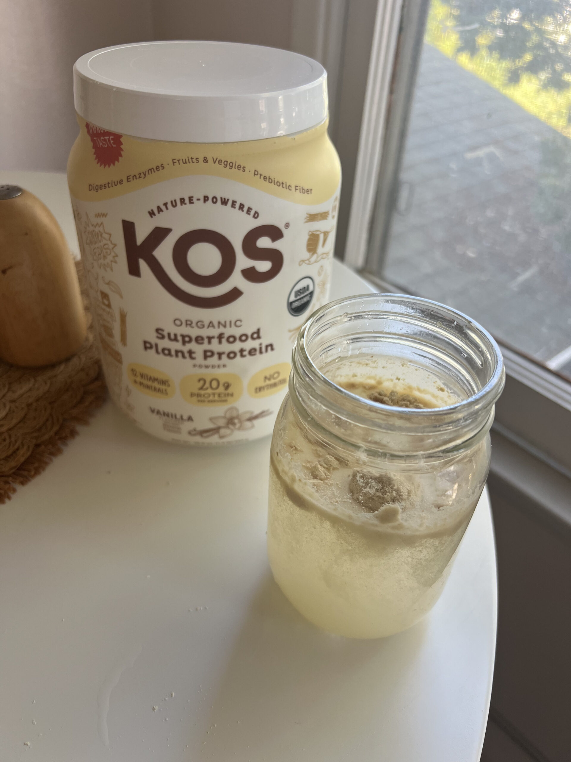 A jar of liquid next to a container of KOS organic superfood plant protein on a table.