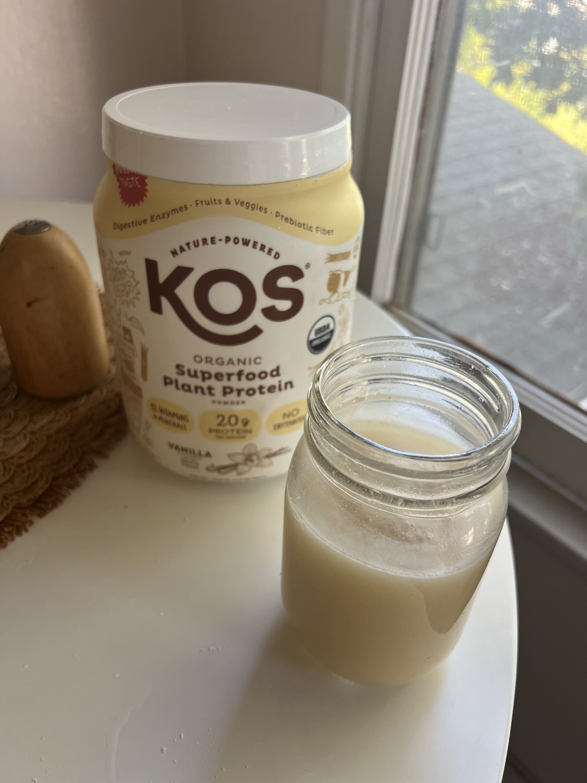 Container of KOS organic plant protein powder and a glass jar with a light-colored liquid on a round white table near a window.