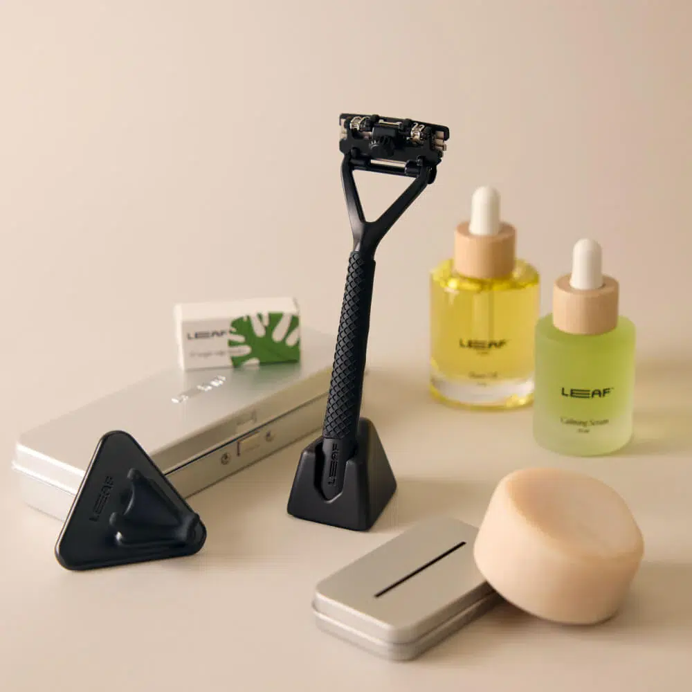 A black razor with a stand is surrounded by skincare bottles, soap, a metal case, and a box.