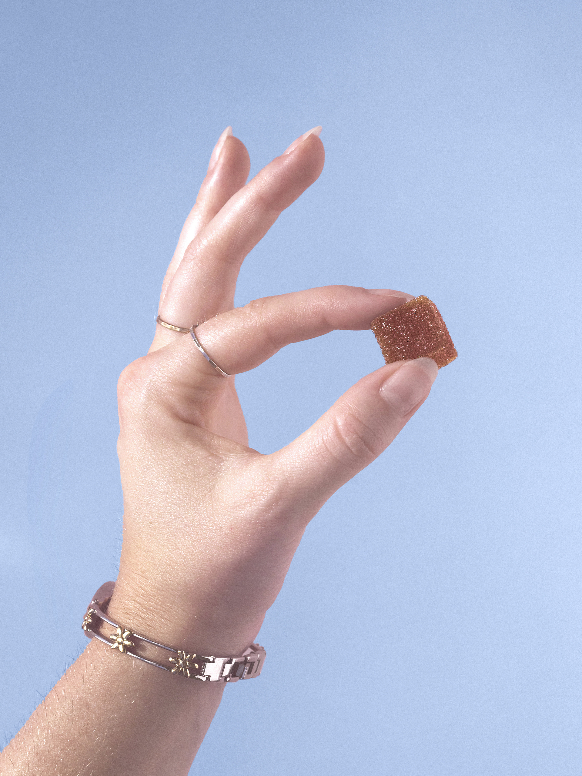 A hand wearing rings and a bracelet holds a small, square, brown gummy candy against a light blue background.