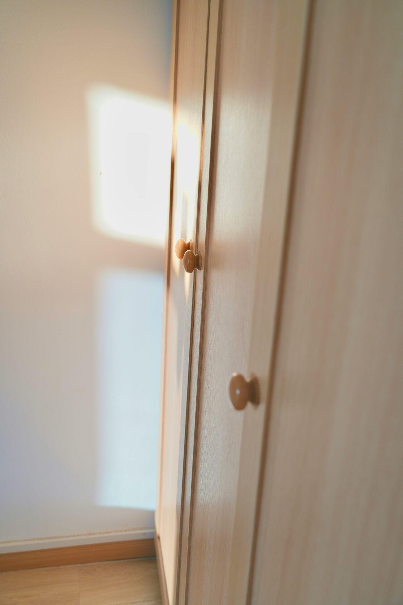 A wooden wardrobe with three doors, each having a round knob, stands against a white wall with sunlight partially illuminating it.