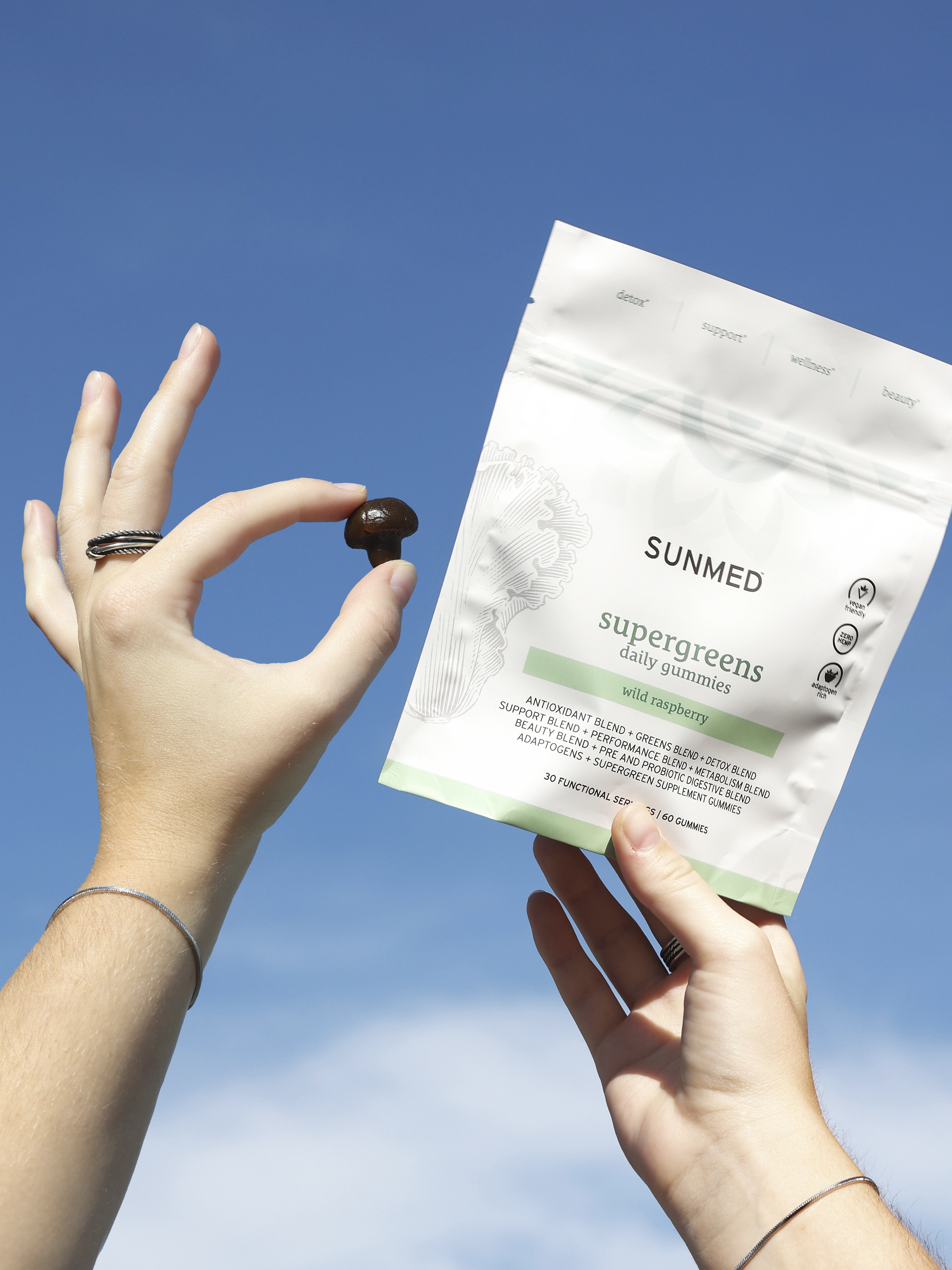 Two hands are shown. The left hand is holding a dark gummy, while the right hand holds a packet of Sunmed supergreens daily gummies against a blue sky background.