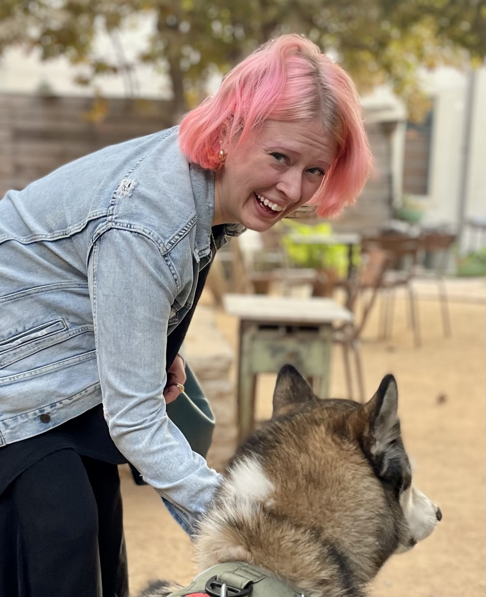 Person with pink hair and denim jacket smiling while petting a large dog outdoors in a park-like setting.