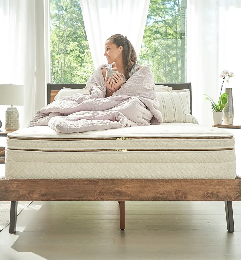 A person wrapped in a light blanket sits on a made bed with an organic mattress, holding a mug, in a brightly lit bedroom with large windows and trees visible outside.