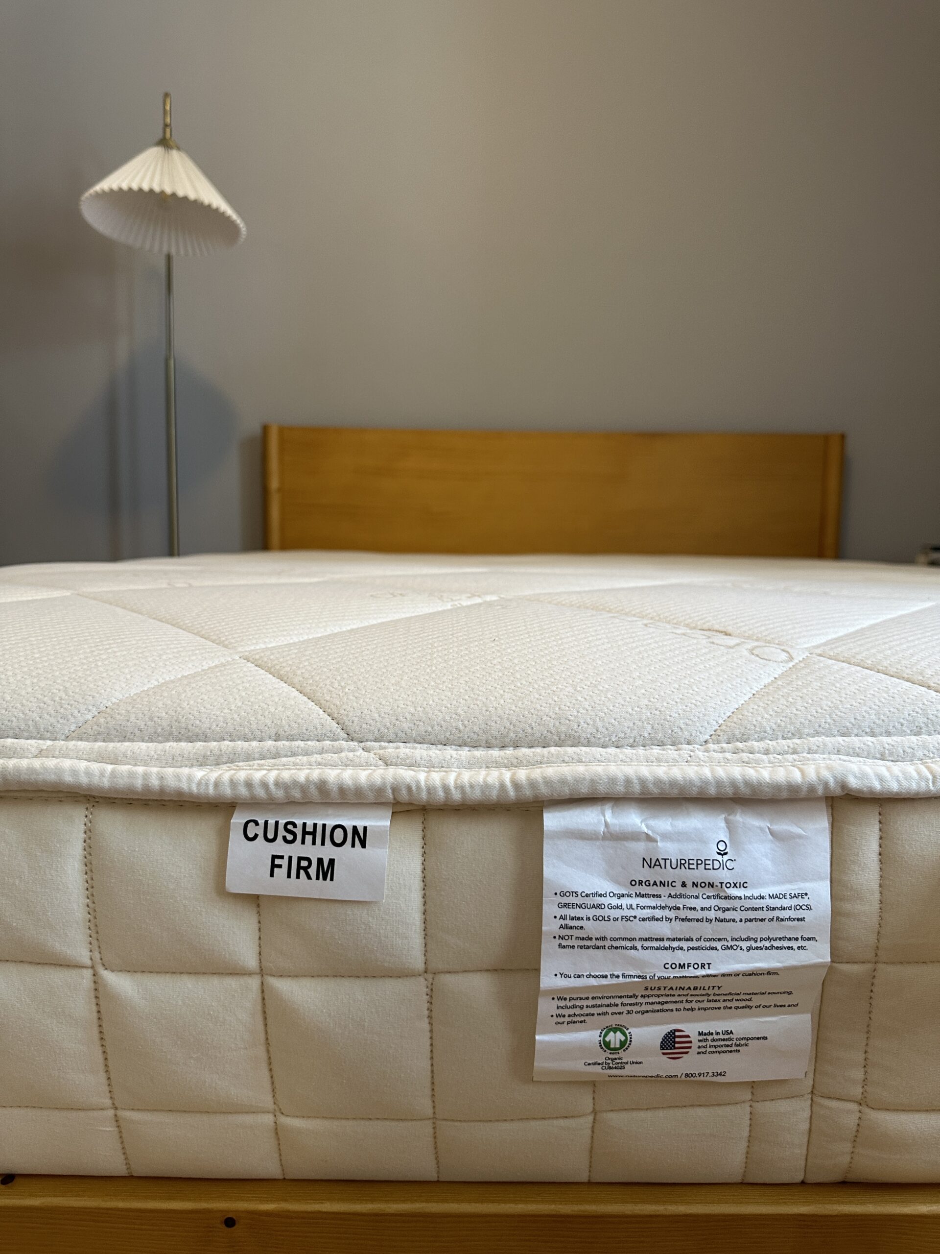 Close-up of a mattress on a bed frame, showing tags labeled "Cushion Firm" and "Naturepedic," with additional care and certification information. A floor lamp is visible in the background.