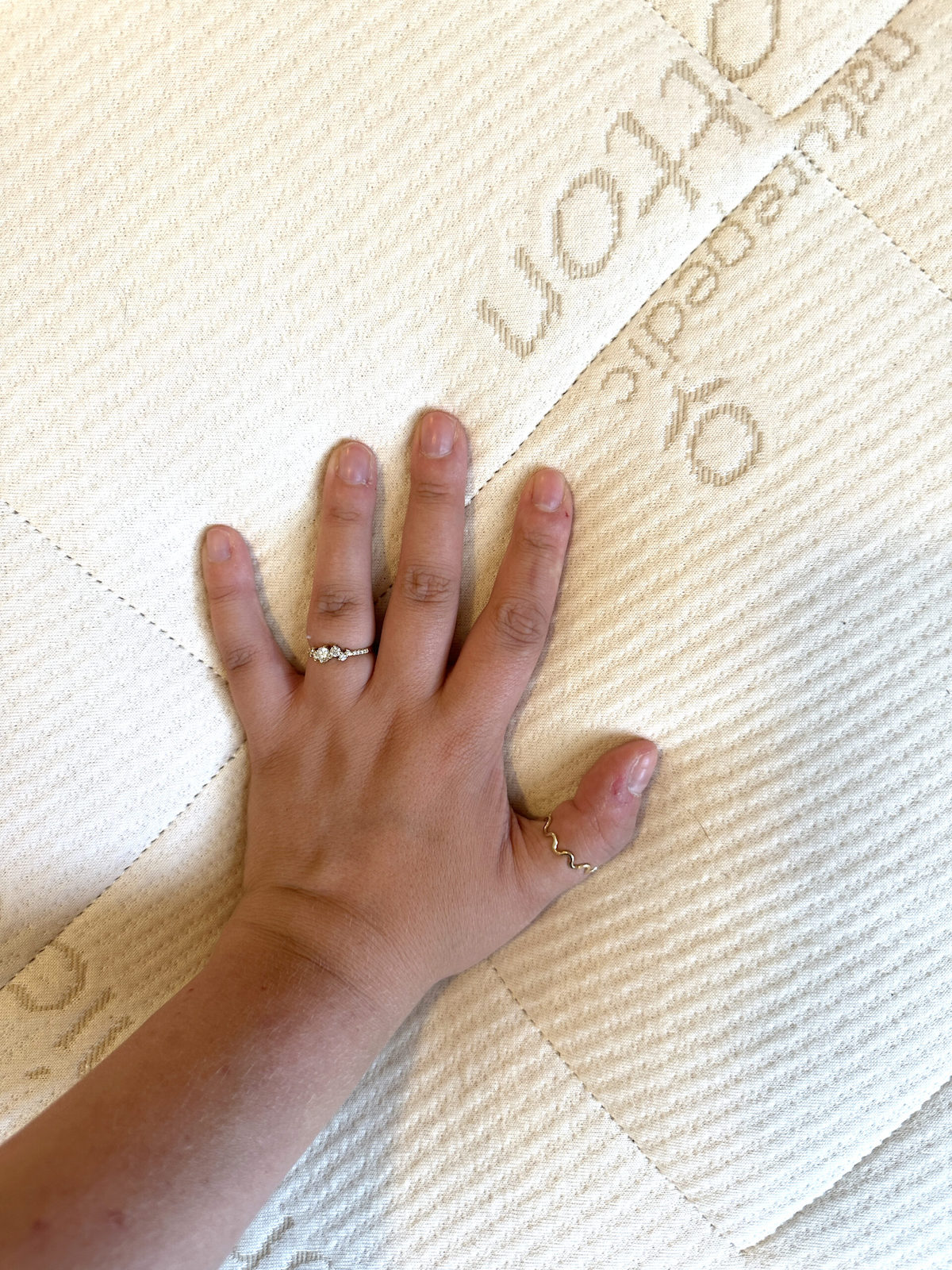 A hand with two rings presses on a textured white surface, possibly an organic mattress or fabric covering.