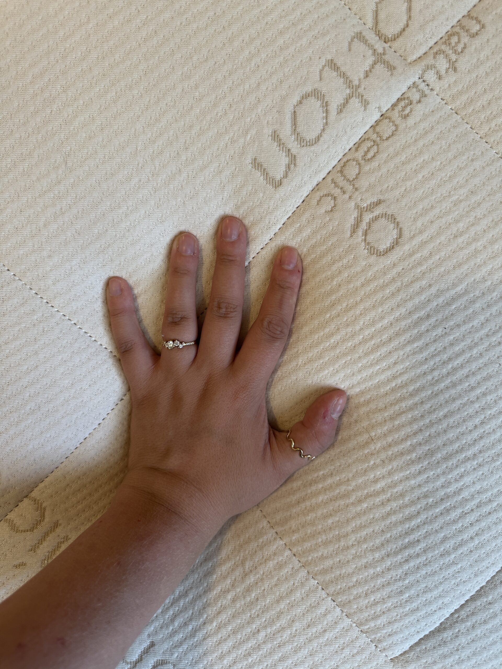 A hand with two rings is placed on a white quilted fabric surface with embossed text.