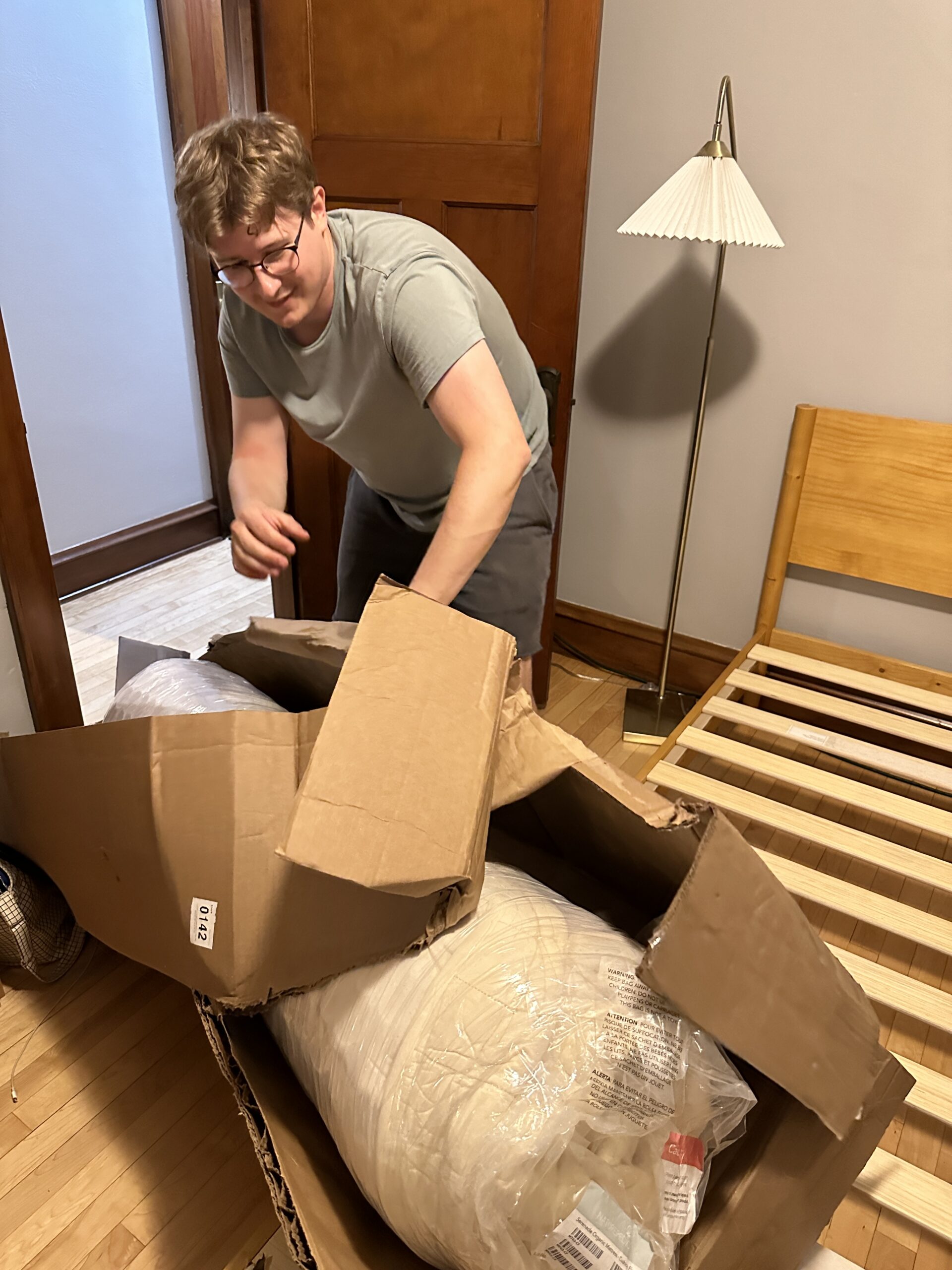 A person wearing glasses and a grey shirt is opening a large, partially damaged cardboard box in a room with wooden floors, a wooden bed frame, and a standing lamp.