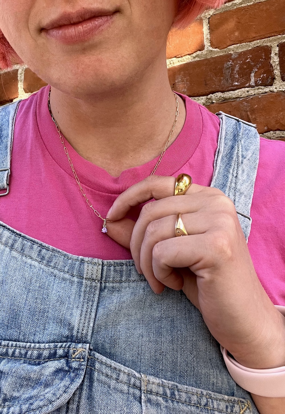 Person wearing a pink shirt and overalls, holding a necklace with a small pendant. They have a gold ring on their finger and stand in front of a brick wall.