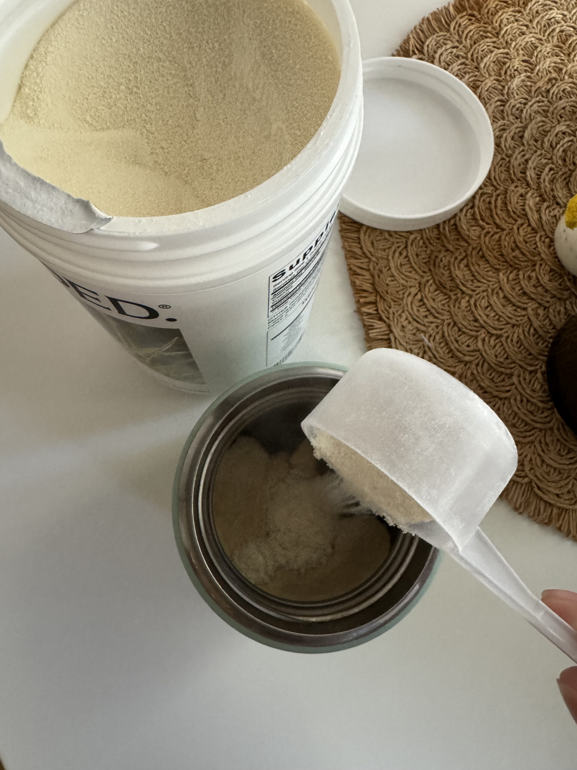 A hand is scooping powder from a large open container into a smaller container on a woven placemat. The scene is set on a white surface.