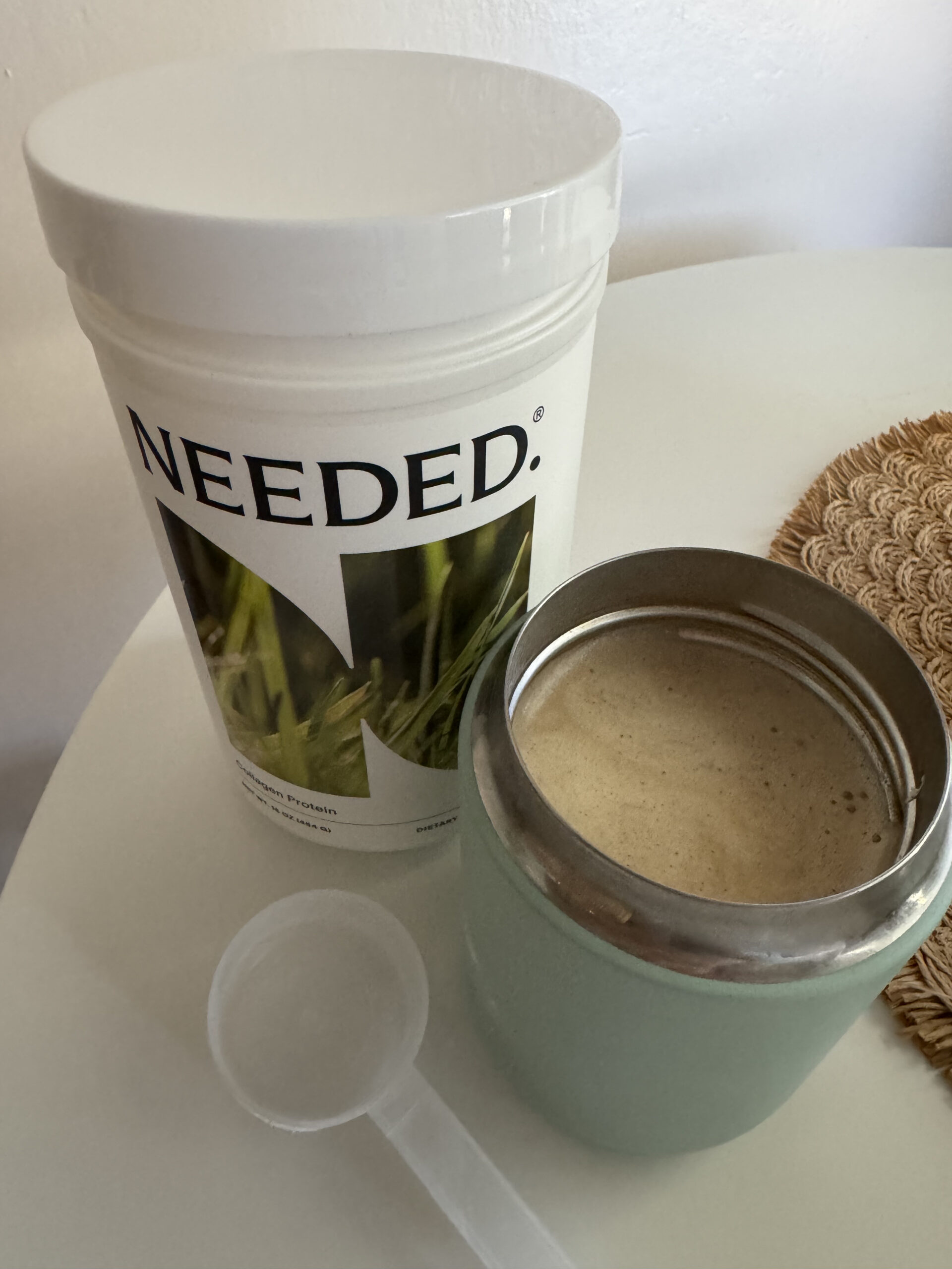 A container labeled "Needed" with a leaf design, next to a filled shaker cup and a plastic scoop on a round table.