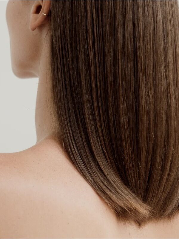 A close-up of a person with straight, shoulder-length brown hair, viewed from behind and slightly to the side, against a neutral background.