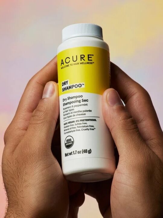 Person holding a bottle of Acure dry shampoo against a soft, multicolored background.