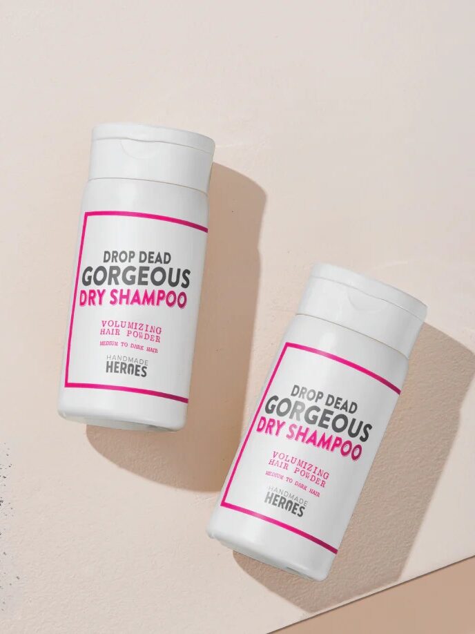 Two bottles of "DROP DEAD GORGEOUS DRY SHAMPOO" lie on a beige surface. One bottle rests horizontally while the other stands upright. A small pile of gray powder is seen next to the bottles.