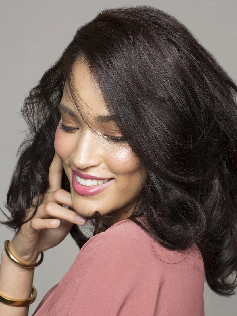 A person with wavy, dark hair and a pink lipstick smiles, touching their face. They are wearing a pink top and gold bangles on their wrist.